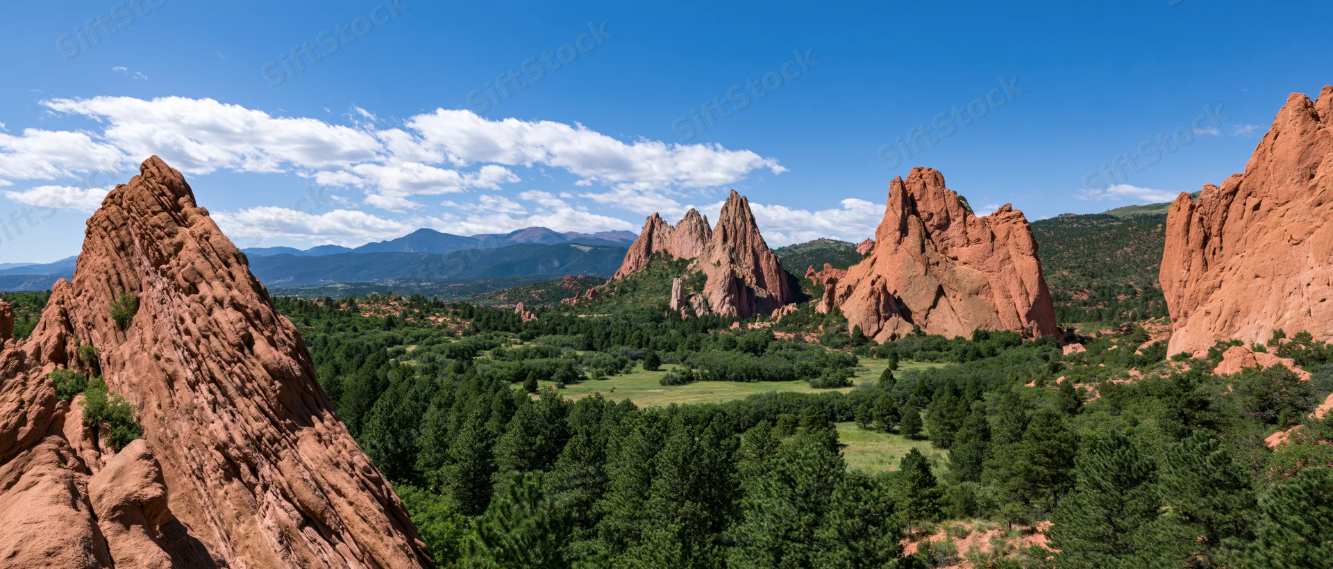 Red sandstone rock formations rising from a green forested valley under a blue sky with scattered clouds