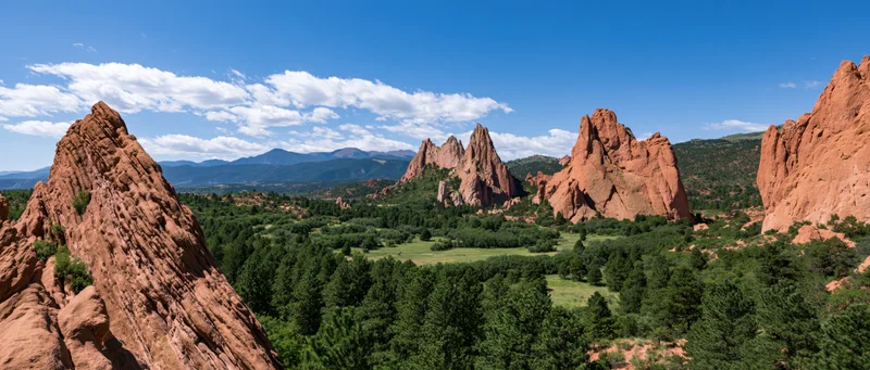Red sandstone rock formations rising from a green forested valley under a blue sky with scattered clouds