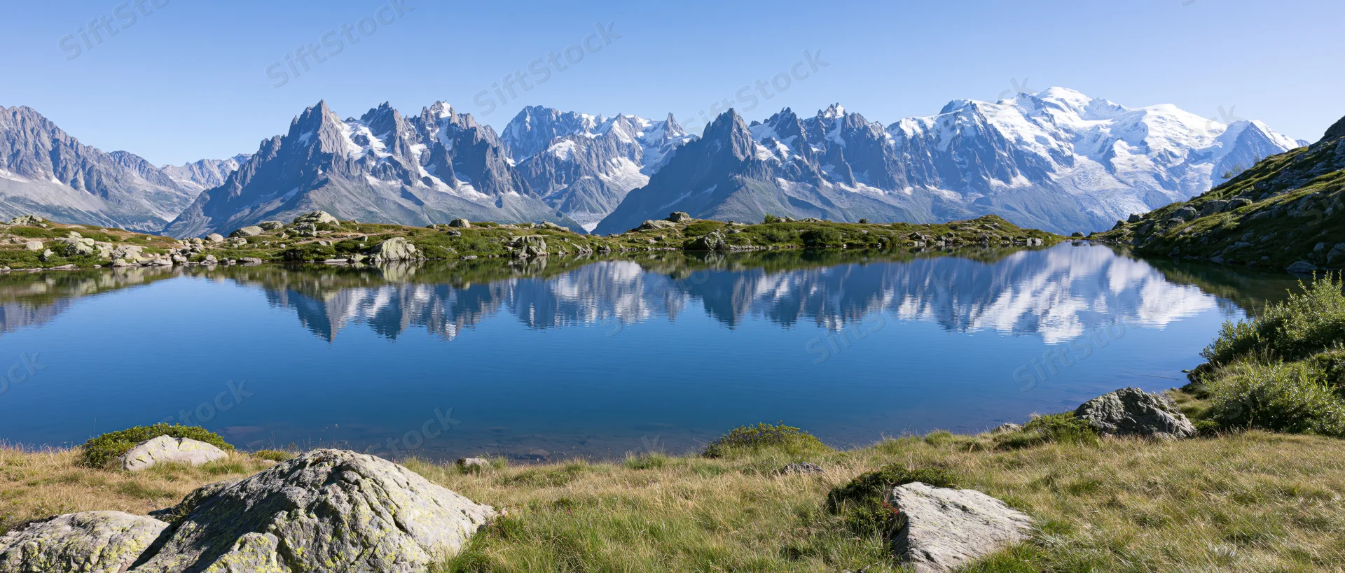 Snow-capped mountain range reflected in calm alpine lake surrounded by grassy meadow and rocks