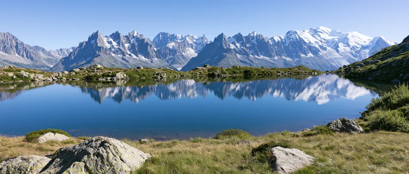Snow-capped mountain range reflected in calm alpine lake surrounded by grassy meadow and rocks