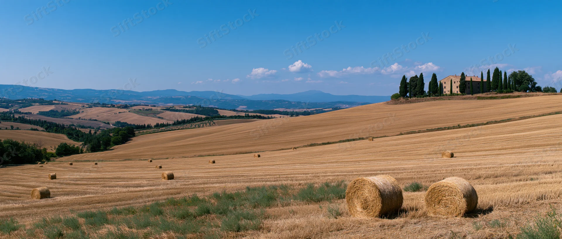 Golden wheat fields with hay bales and a stone villa on a hill in tuscany under a blue sky