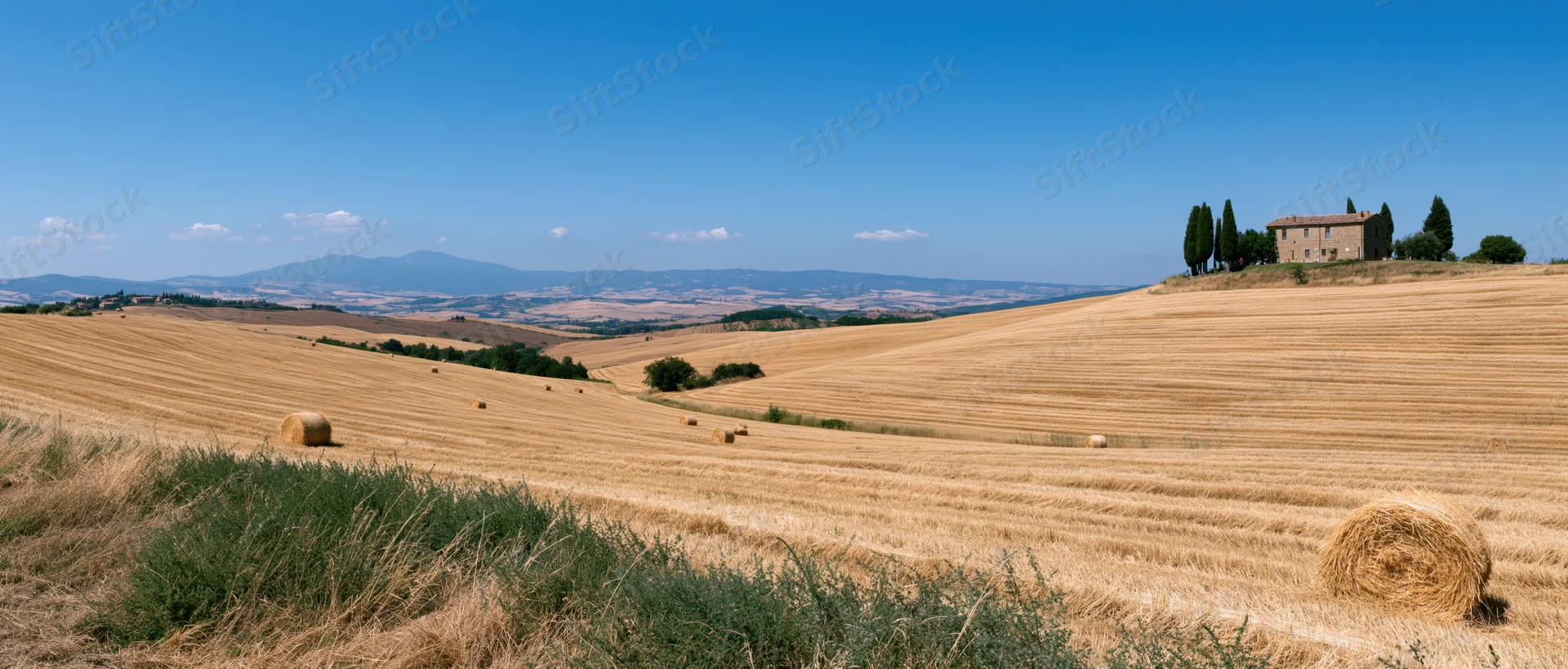 Golden wheat fields with haybales and a stone farmhouse on rolling hills under a clear blue sky