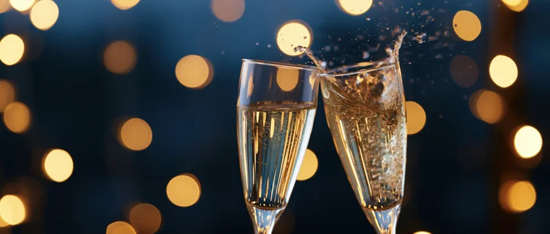 Two champagne glasses clinking with splashing liquid against a dark background with warm bokeh lights