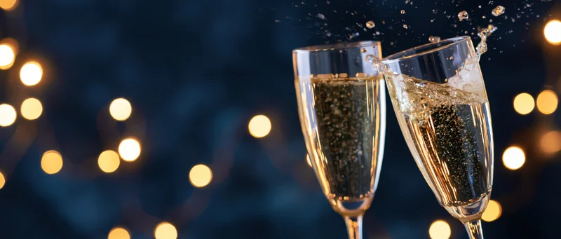 Two champagne glasses clinking with bubbles splashing against a dark background with warm bokeh lights