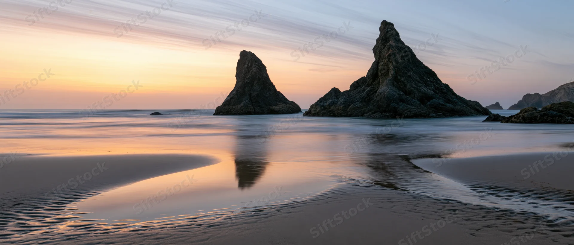 Rocky sea stacks reflected in wet sand at sunset on a calm beach