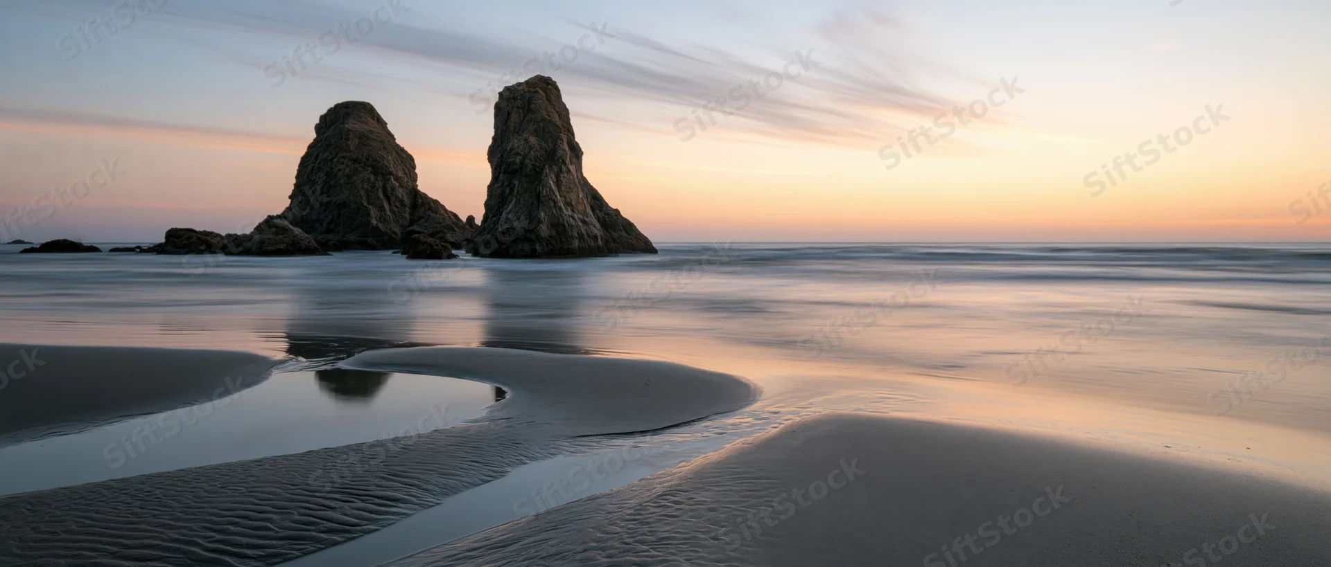 Two large sea stacks rising from the ocean at sunset on a smooth sandy beach with tide pools and reflections
