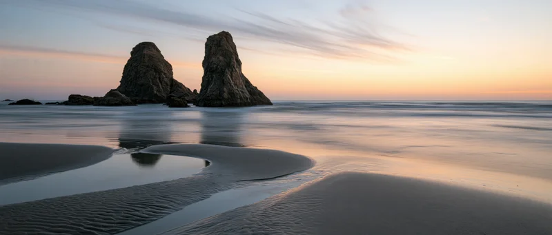 Two large sea stacks rising from the ocean at sunset on a smooth sandy beach with tide pools and reflections