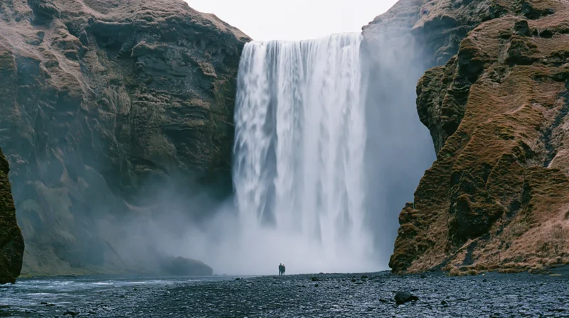 A lone person standing before a massive waterfall cascading between rocky cliffs in a misty natural landscape