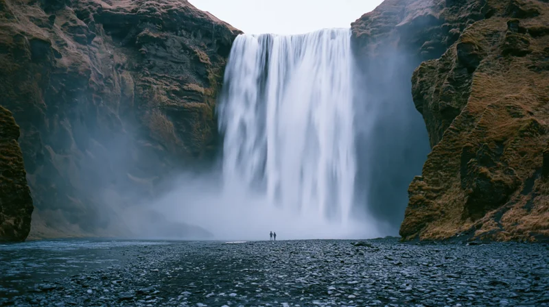 Waterfall cascading down rocky cliffs into a misty river with two people standing at the base