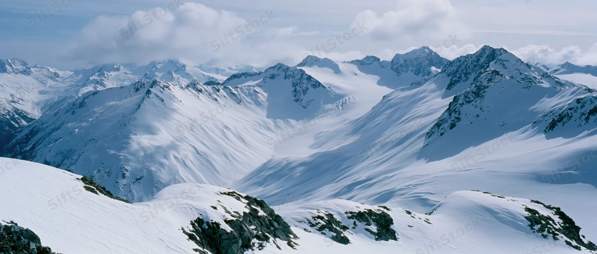 Snow-covered mountain peaks and ridges forming a vast alpine landscape under a cloudy sky