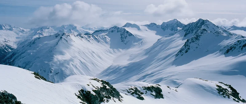 Snow-covered mountain peaks and ridges forming a vast alpine landscape under a cloudy sky
