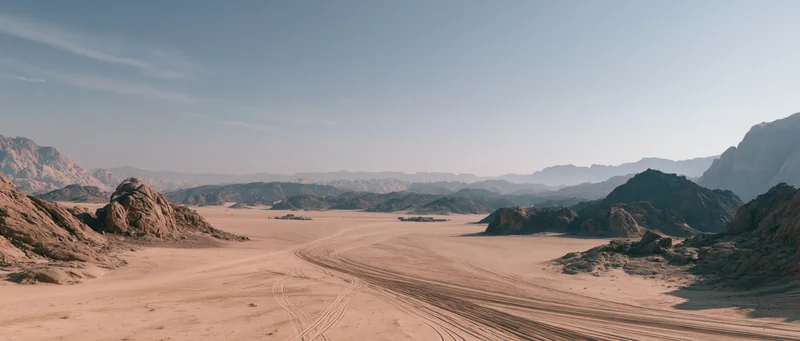 Vast desert valley with tire tracks winding through sandy terrain between rocky mountains under clear blue sky