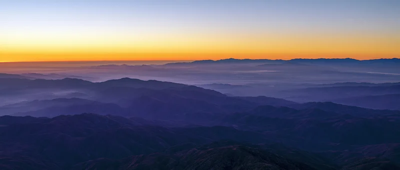 Layered mountain ranges under a gradient sunset sky with fog in the valleys