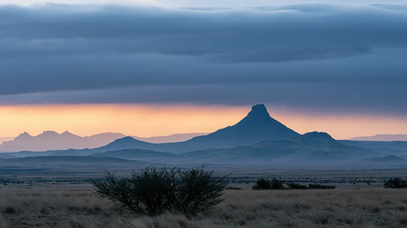 Silhouetted mountain formation at sunset over arid desert landscape with sparse scrub and distant ridges