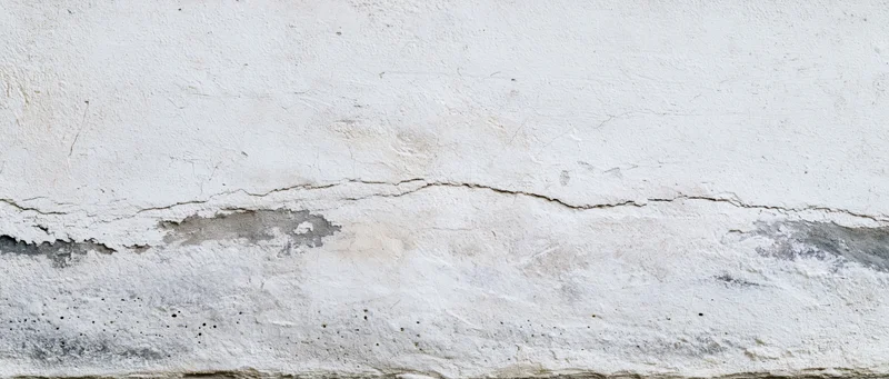 Cracked and damaged white plaster wall showing concrete foundation beneath