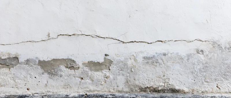 Cracked white plaster wall showing structural damage at base near floor