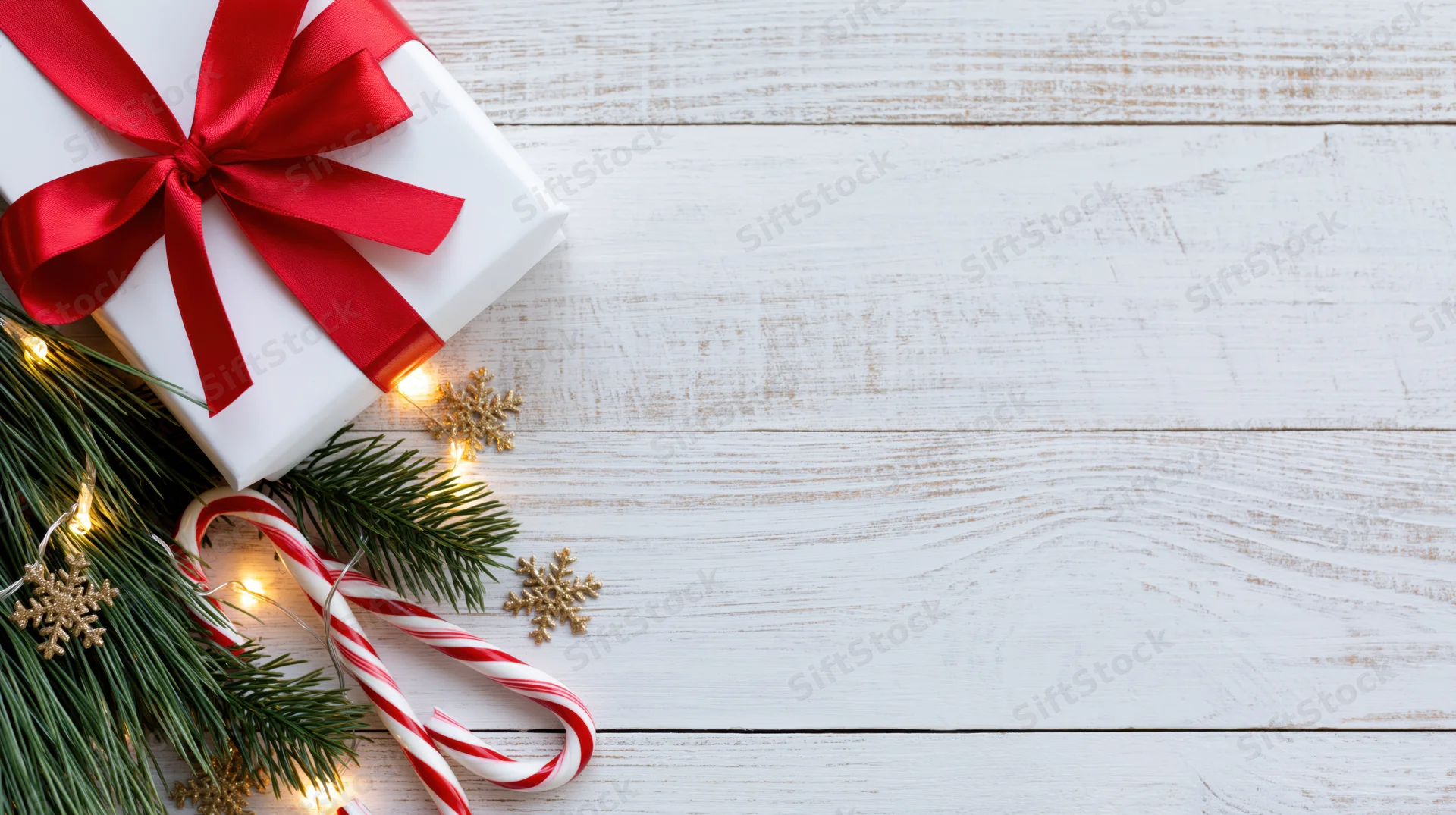 White gift box with red ribbon and candy canes on wooden surface with pine branches and lights