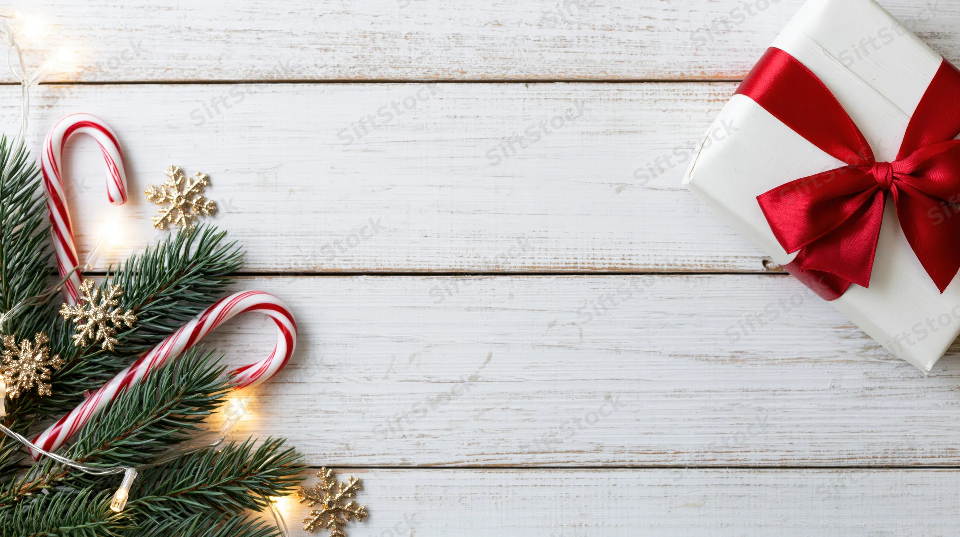 Christmas gift wrapped in white paper with red ribbon beside pine branches, candy canes, and gold snowflakes on a white wooden surface