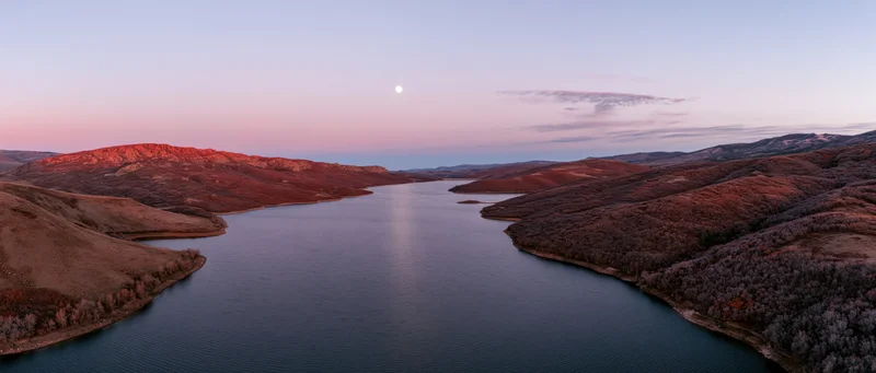 Aerial view of a calm lake surrounded by rolling hills and forests at twilight with moon in sky