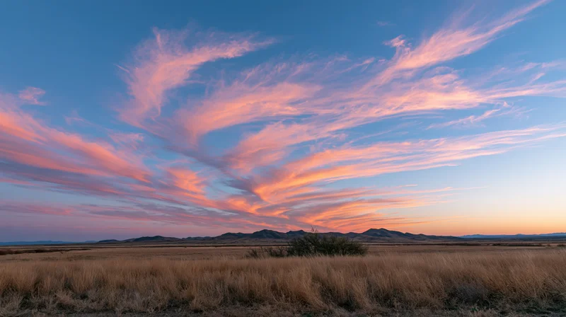 Pink and orange clouds over a desert grassland at sunset with distant mountains on the horizon