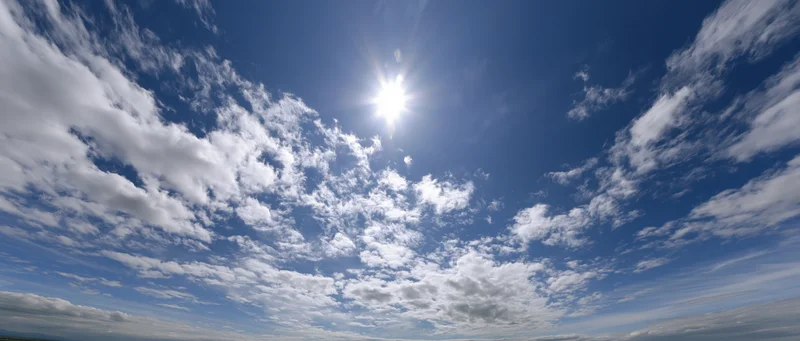 Bright sun shining through scattered white clouds in a deep blue sky with visible horizon line
