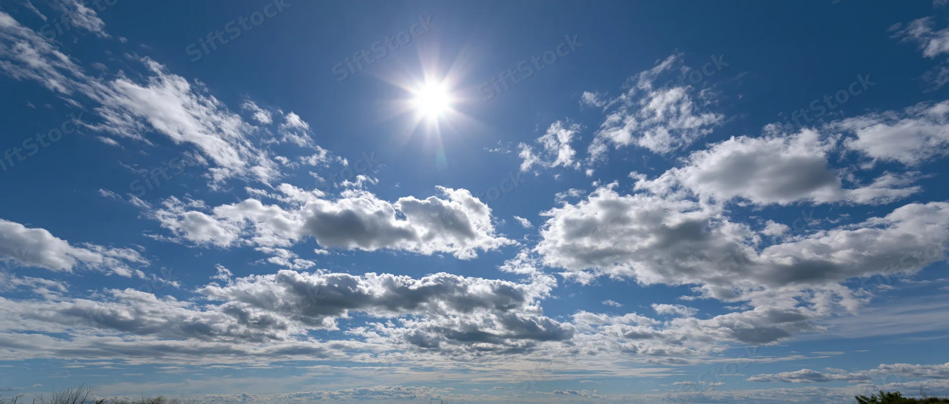 Bright sun shining through scattered white clouds in a clear blue sky above open landscape
