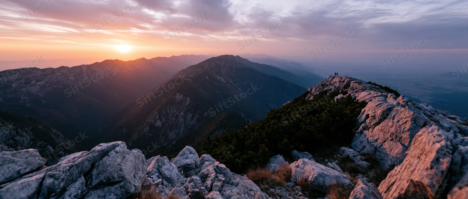 Rocky mountain peak at sunset overlooking a valley with distant ridges and a hiker on the summit