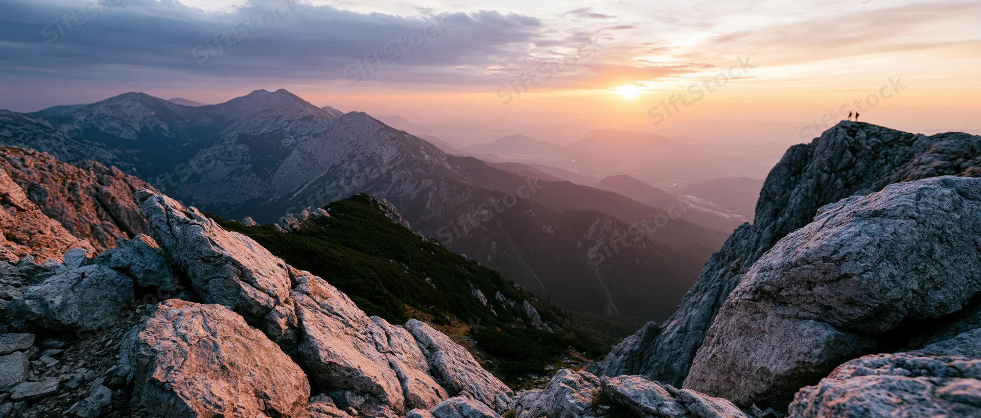 Two hikers standing on a rocky mountain peak overlooking a vast valley at sunset