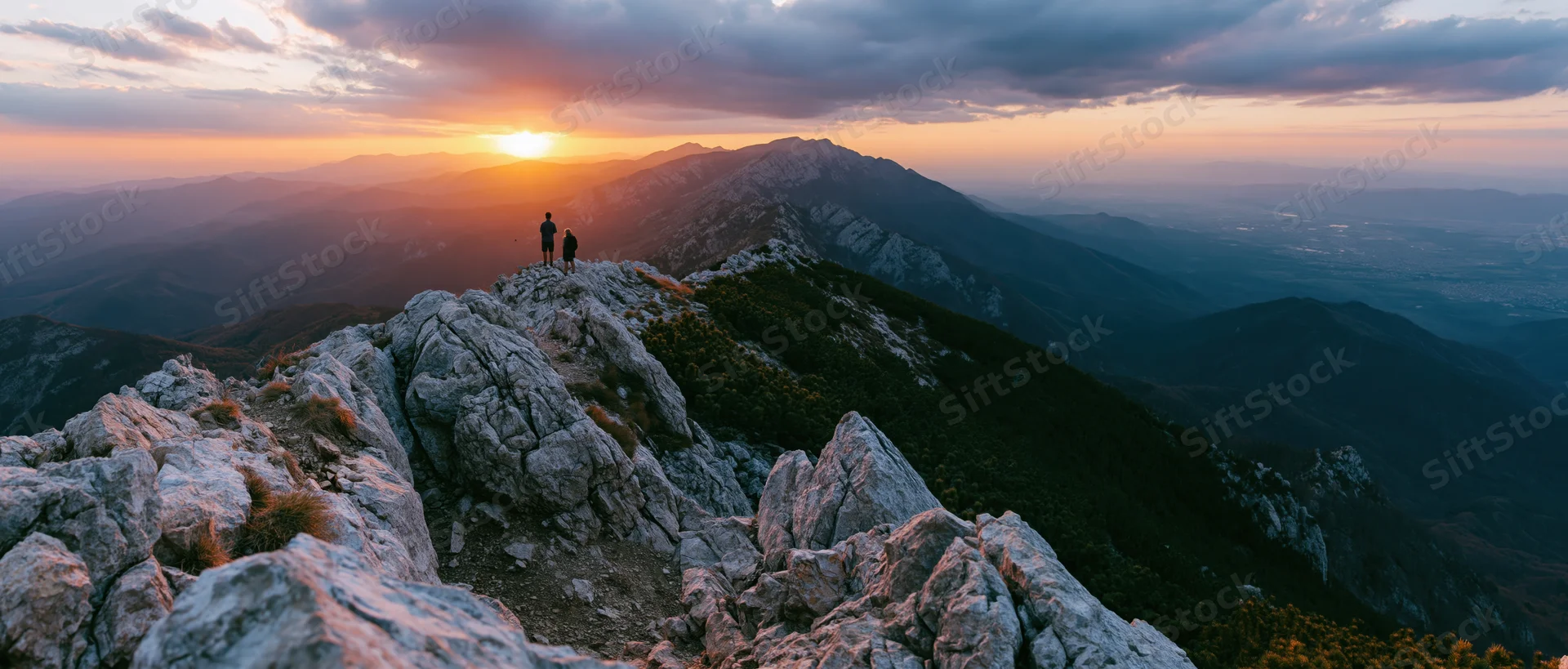 Couple standing on rocky mountain peak at sunset overlooking valley and distant mountains