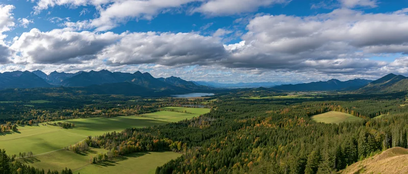 Panoramic view of a mountain valley with forests and meadows under a cloudy blue sky in autumn