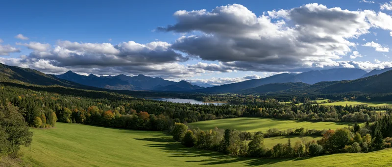 Panoramic view of a green valley with forested hills and a lake under a cloudy sky