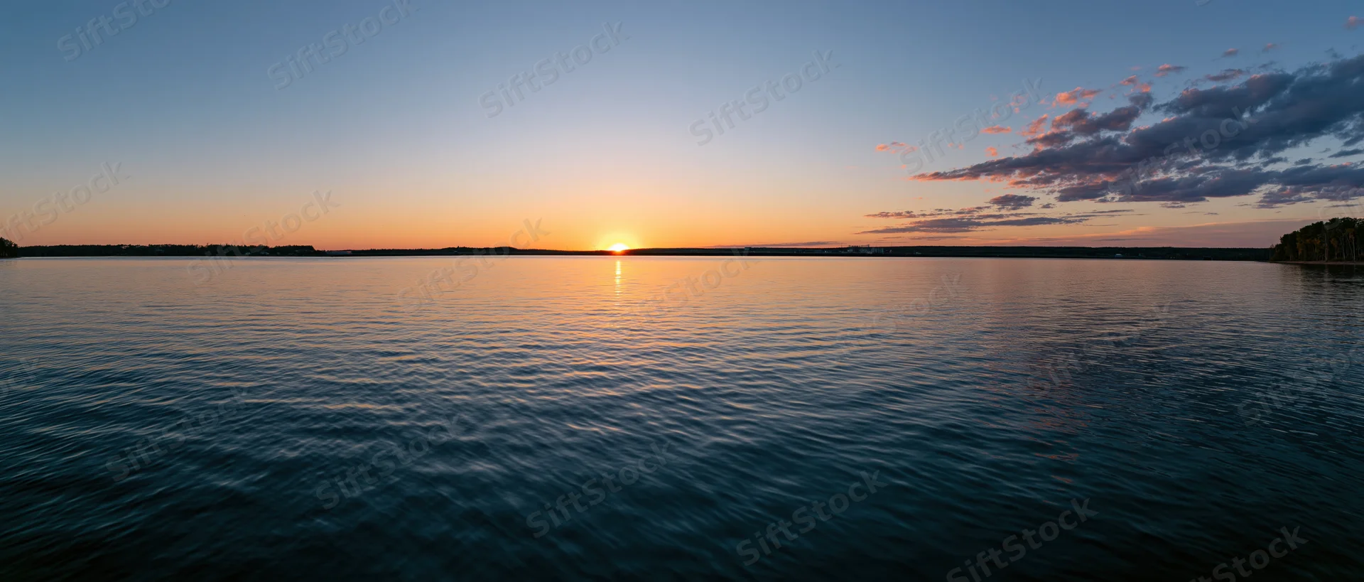 Sunset over calm lake reflecting golden light on rippled water with distant tree-lined shore under twilight sky
