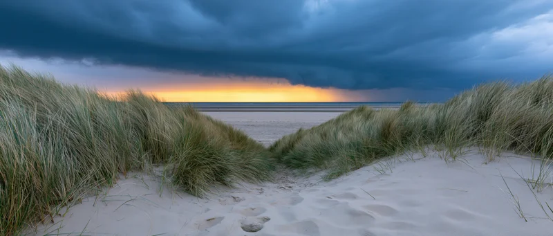 Sandy path between dunes leading to ocean under stormy sky at sunset