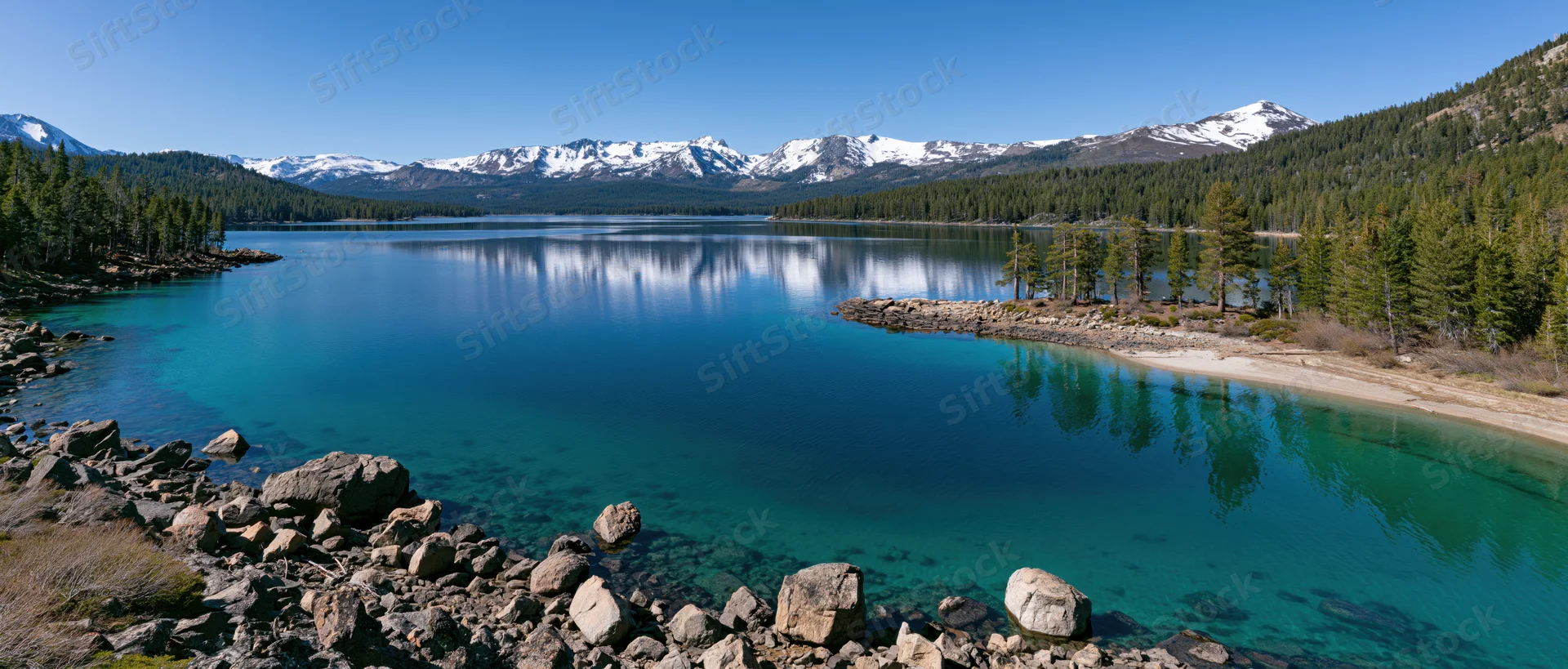 Calm turquoise lake reflecting snow-capped mountains and pine forest along rocky shore