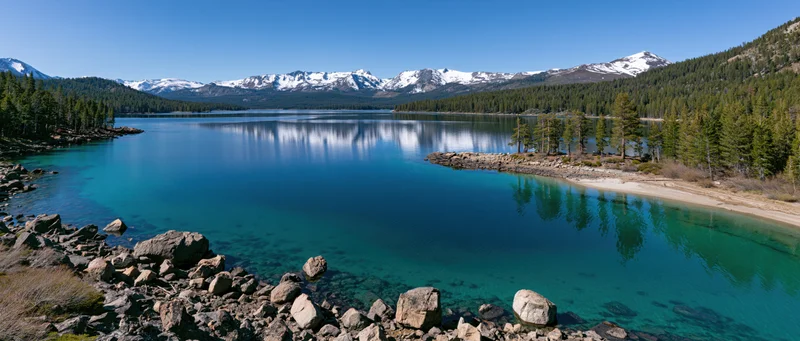 Calm turquoise lake reflecting snow-capped mountains and pine forest along rocky shore