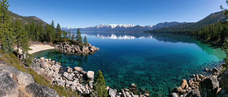Calm alpine lake reflecting snow-capped mountains surrounded by pine forest and rocky shore