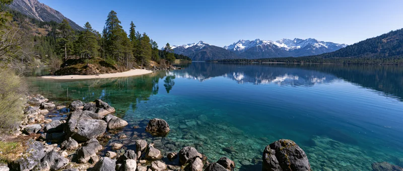 Calm alpine lake reflecting snow-capped mountains and forested shore under clear blue sky