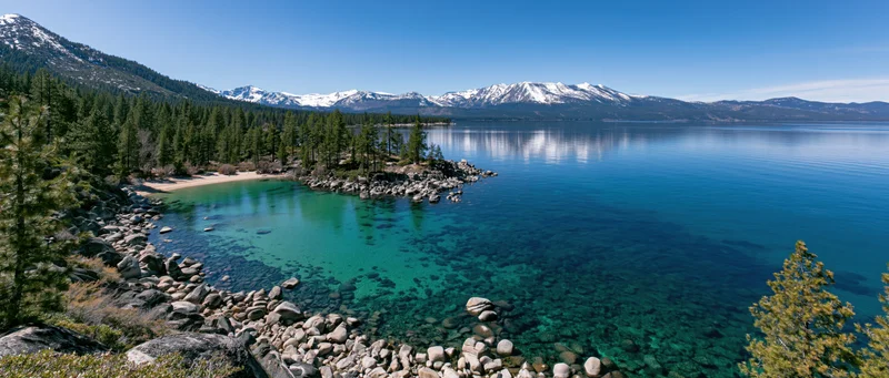 Calm turquoise lake bordered by rocky shore and pine forest with snow-capped mountains in the distance