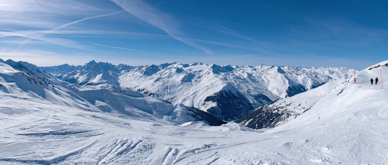 Snow-covered alpine mountains with ski slopes and distant skiers under a clear blue sky