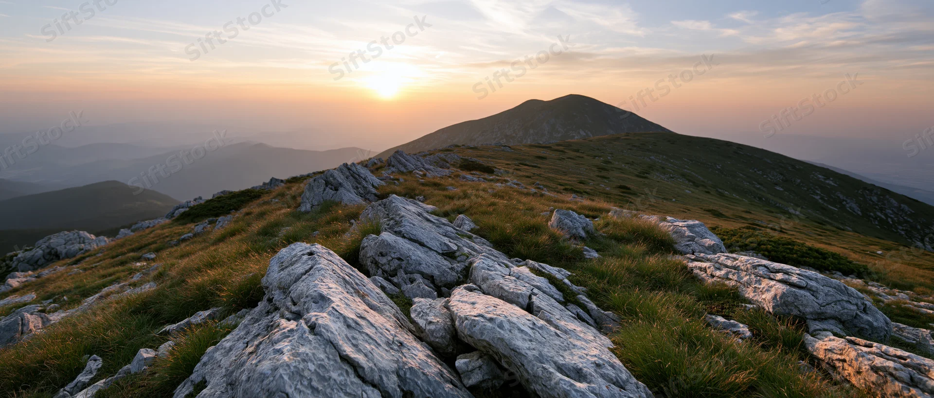 Rocky mountain ridge at sunset with grassy slopes and distant hills under golden sky