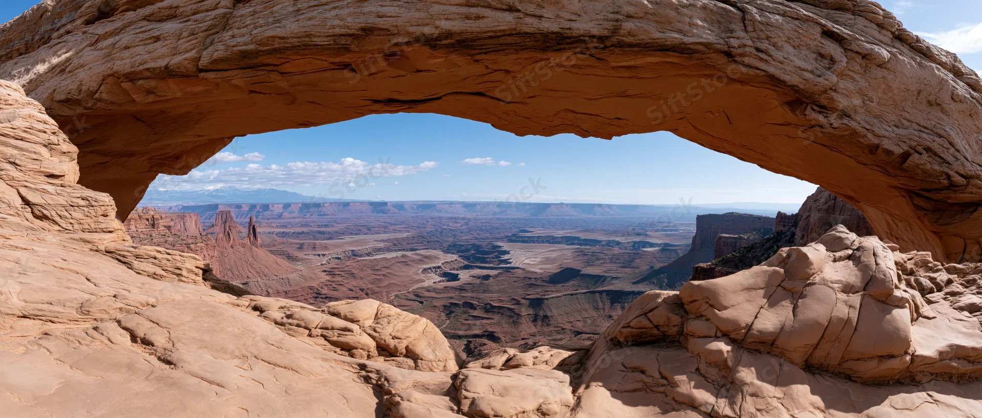 Natural sandstone arch framing a vast desert canyon landscape under a blue sky with scattered clouds