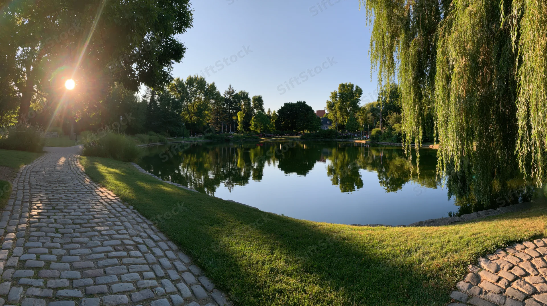 Cobblestone pathway winding beside a calm pond with willow trees and green grass in a park at morning sunlight
