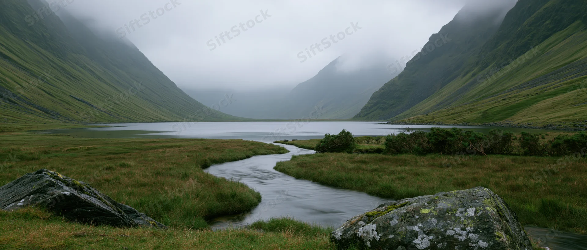Winding river flows through grassy valley toward misty mountains in scottish highlands