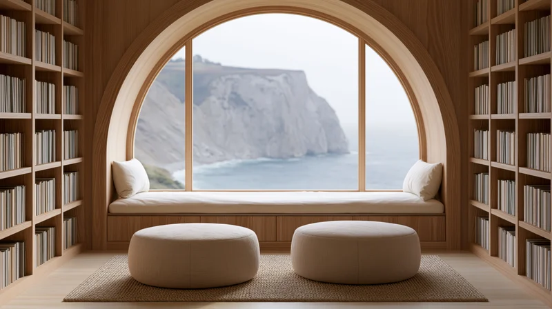 Wooden reading nook with window seat overlooking ocean cliffs, flanked by bookshelves and furnished with cushions and ottomans