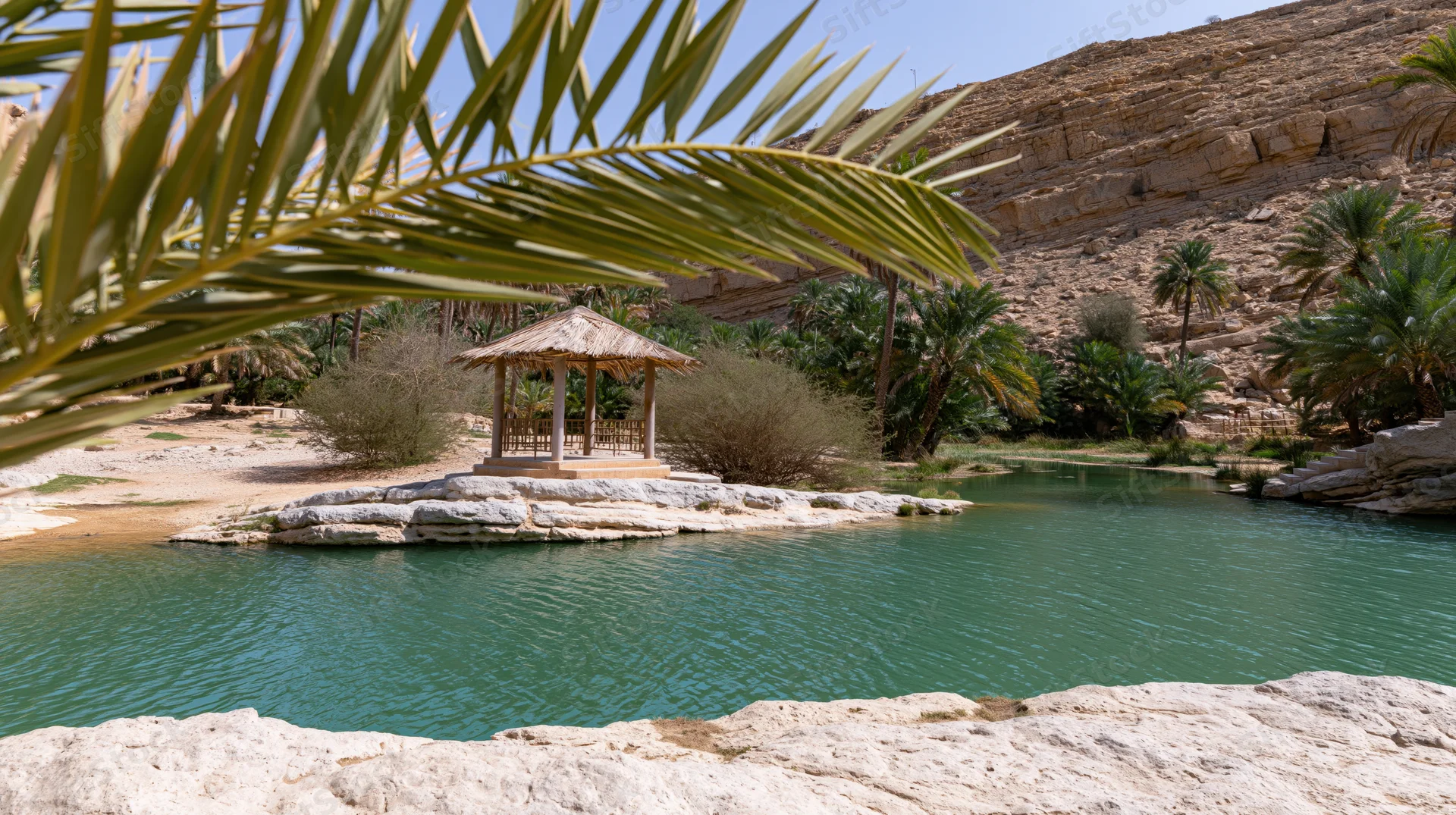Thatched hut on a rocky island in a green pool surrounded by palm trees and desert cliffs