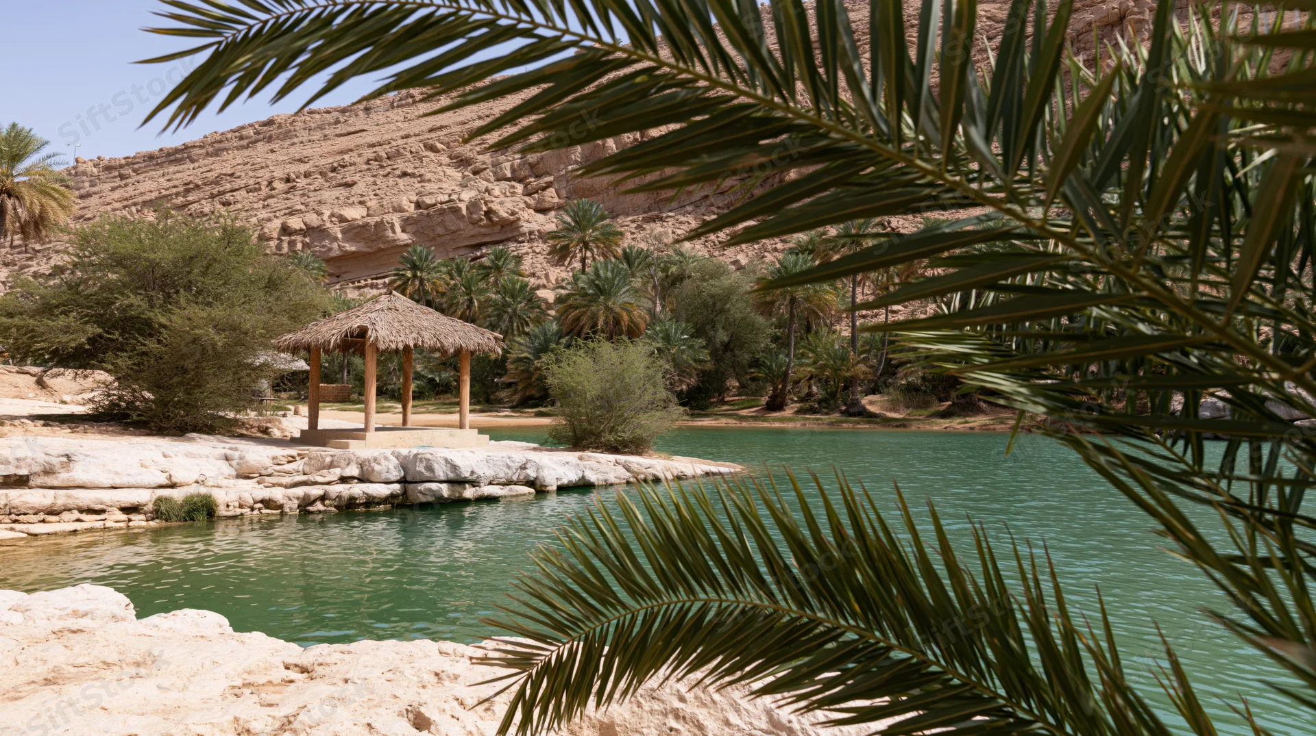 Palm fronds framing a tranquil oasis with a thatched hut by turquoise water in a desert landscape