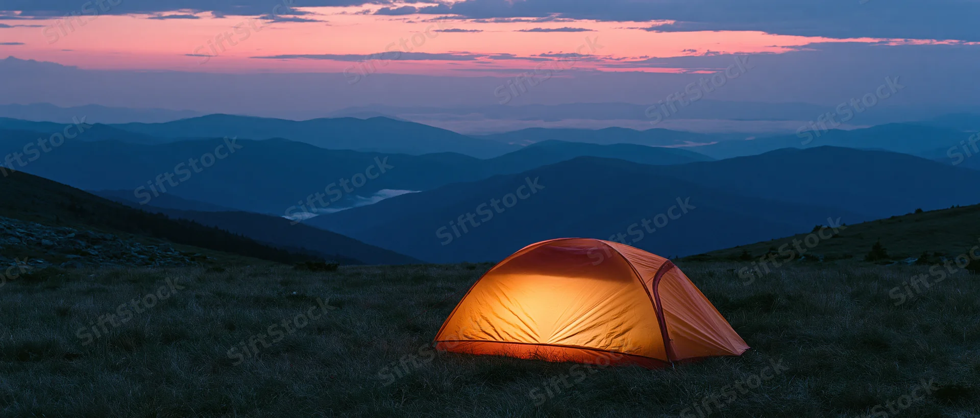 Illuminated tent on grassy mountain ridge at twilight overlooking layered valleys