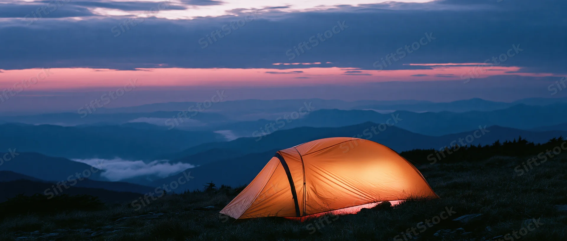 Glowing tent on mountain ridge at twilight overlooking layered valleys and distant peaks