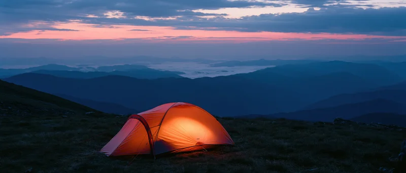 Illuminated tent on mountain summit at twilight overlooking layered valleys and clouds