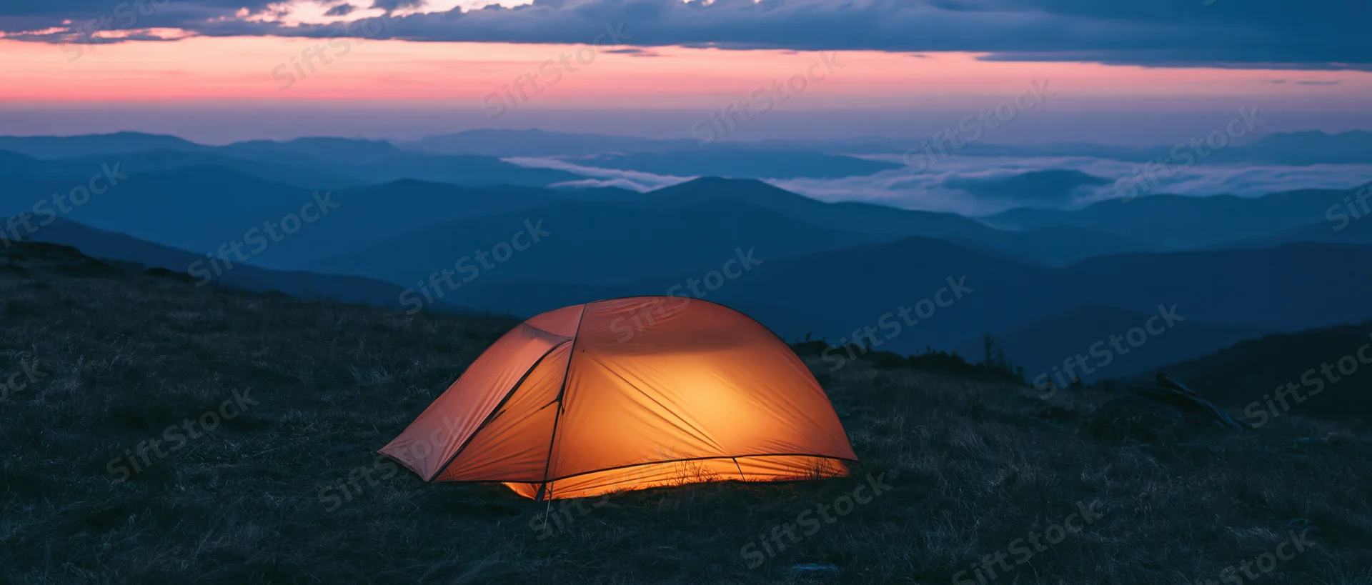 Illuminated tent on grassy mountain ridge at twilight overlooking layered valleys and fog under pink and blue sky